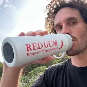 White insulated Red Gum Property Management drink bottle with red logo, held outdoors
