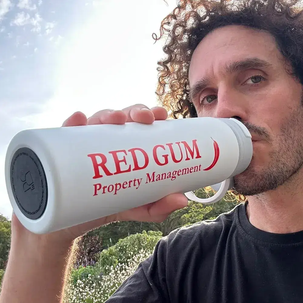 White insulated Red Gum Property Management drink bottle with red logo, held outdoors